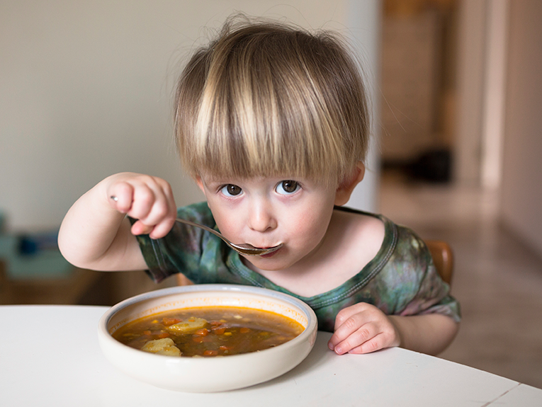 A young child with blonde hair and brown eyes sits at a kitchen table eating soup