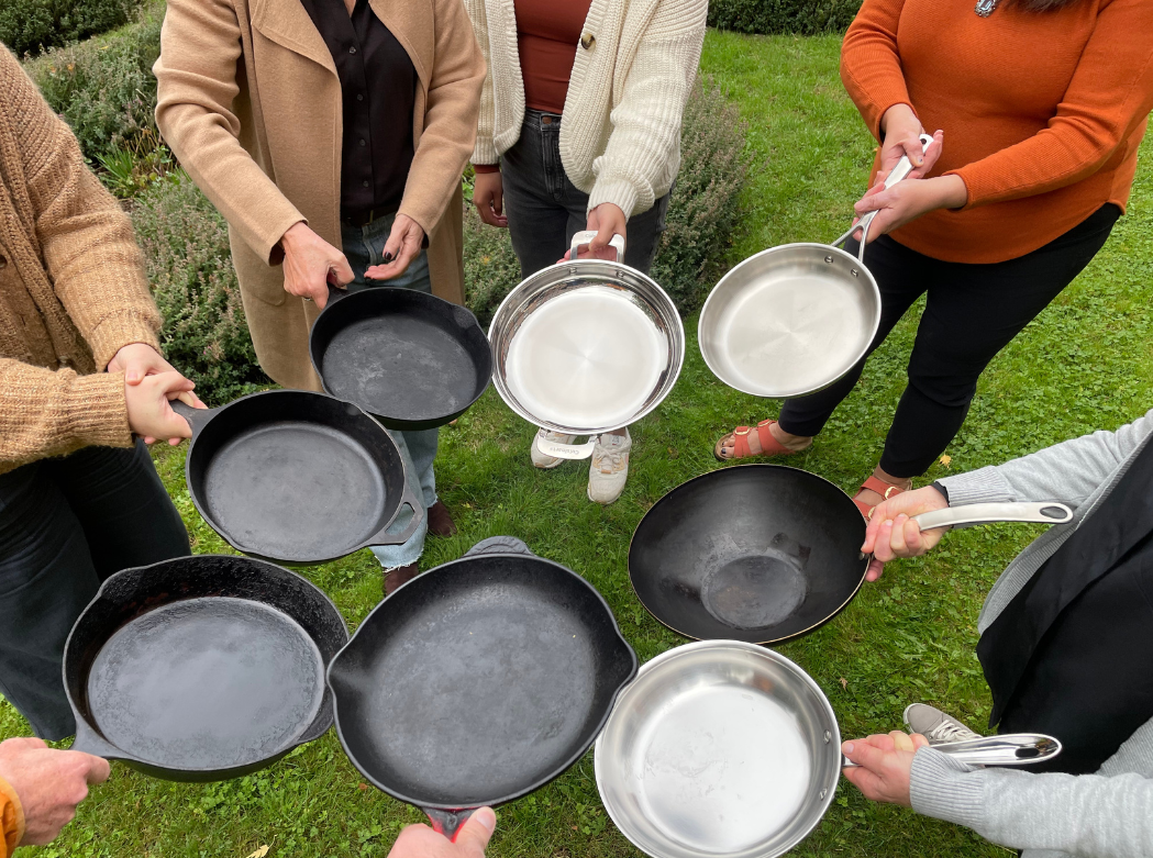 A group of people standing in a circle, holding stainless steel and cast iron pans.