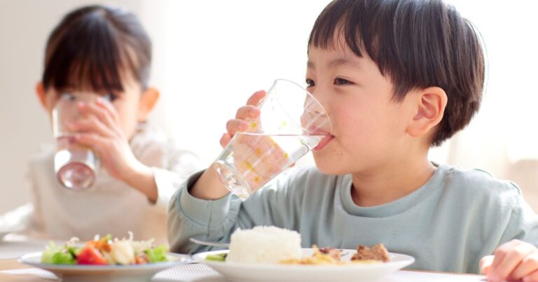 Two children sitting at the table with plates of food, drinking glasses of water.