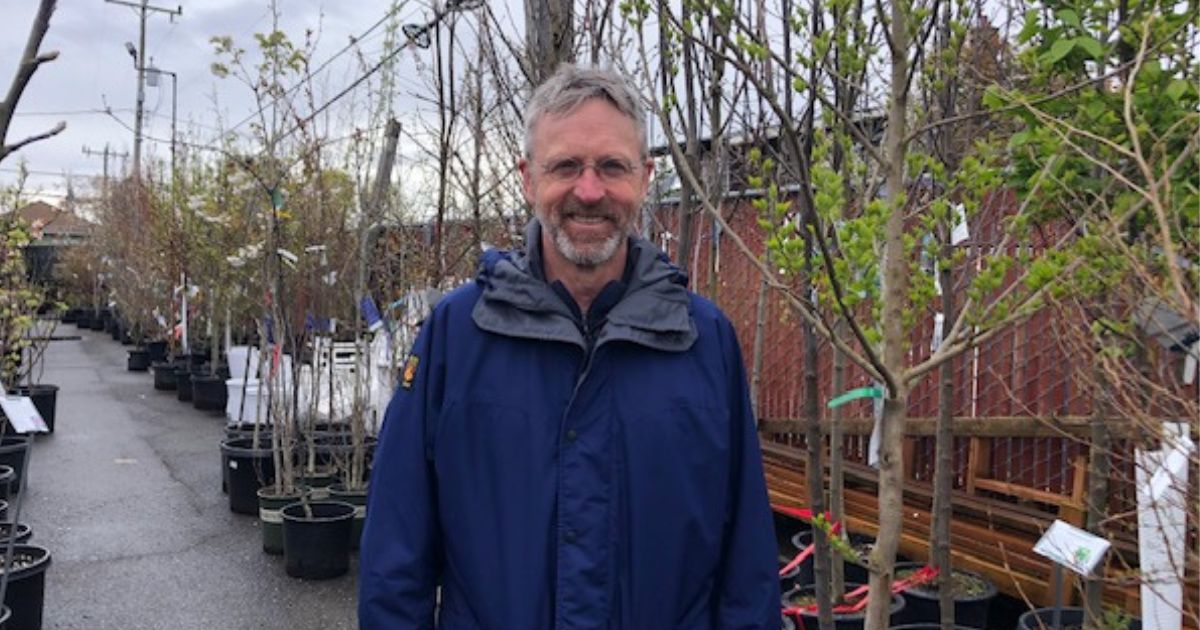 A man wearing a blue coat, standing in a garden center in front of some plants